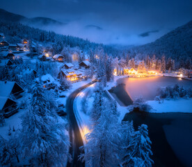 Aerial view of fairy town in snow, road, forest, Jasna lake and houses with lights at night in winter. Top view of mountain village, illumination, snowy pine trees at dusk in Kranjska Gora, Slovenia - obrazy, fototapety, plakaty