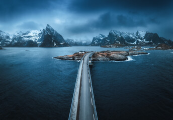 Aerial view of bridge, island with rorbu, sea, snowy mountains in fog, blue cloudy sky at twilight in spring. Dramatic landscape. Top drone view of road. Hamnoy village, Lofoten islands, Norway