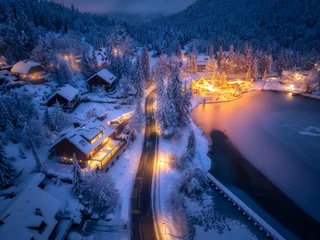 Fotobehang Aerial view of fairy town in snow, road, forest, Jasna lake and houses with lights at night in winter. Top view of mountain village, illumination, snowy pine trees at dusk in Kranjska Gora, Slovenia © den-belitsky