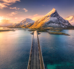 Aerial view of bridge over the sea and snowy mountains in Lofoten Islands, Norway. Fredvang bridges at sunset in winter. Landscape with blue water, rocks in snow, road, sky with clouds. Top drone view