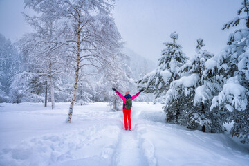 Happy young woman with backpack and raised up arms in fairy snowy forest in winter. Kranjska Gora, Slovenia. Trees in snow, hoar, sporty girl, trail. Snowfall in foggy woods. Wintry woodland. Hiking 