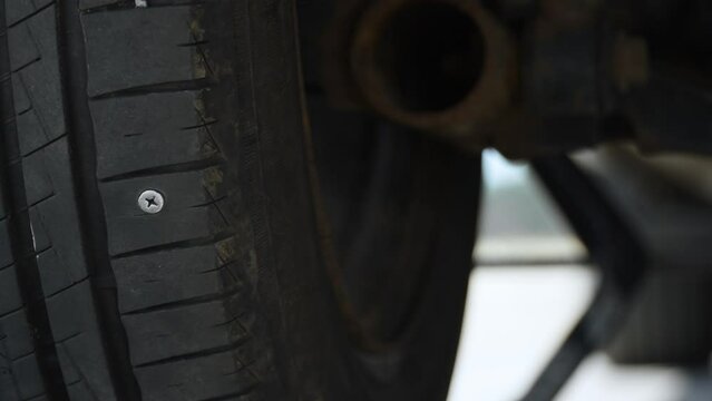 Screw appears on rotating damaged tire of jacked up car, puncturing wheel. Car mechanic touches and checks condition of tire tread with his hand wearing protective gloves.