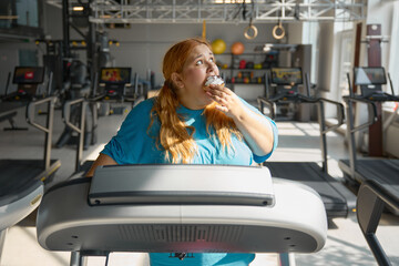 Front view on funny obese woman eating cake while training on treadmill at gym