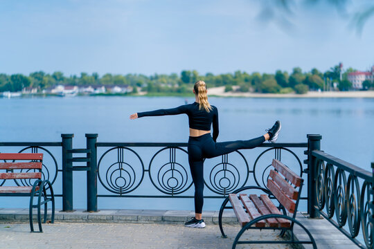 A girl athlete does morning stretching for leg flexibility on the embankment overlooking river