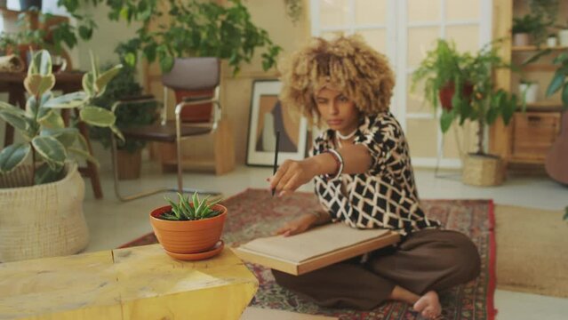 Medium long shot of African American woman painting on canvas from potted houseplant while sitting in lotus position at home surrounded by plants