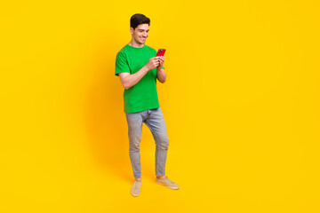 Full size photo of good mood guy wear stylish t-shirt denim pants look at smartphone read notification isolated on yellow color background