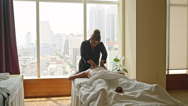 Mulracial masseuse gently massages back of a young woman at luxury spa center with a stunning view. Biracial spa center female employee performs wellness procedure to client at recreation facility