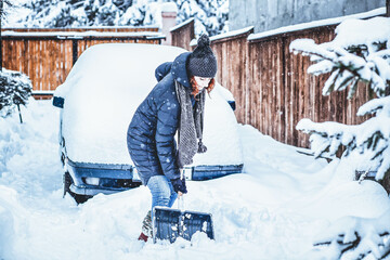woman with shovel cleaning snow around car. Winter shoveling. Removing snow after blizzard - obrazy, fototapety, plakaty