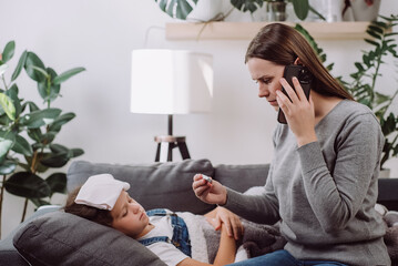 Sad sick preteen kid girl lying on sofa feel fever catch cold flu influenza. Anxious mom holding phone to ear make call to doc family therapist after measuring child temperature. Calling family doctor - Powered by Adobe