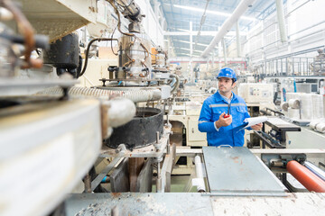 American male engineer, technician manager Standing and looking at the working machinery In the plastic and steel industry, holding a list of notes and a radio, wearing a uniform and a safety helmet.