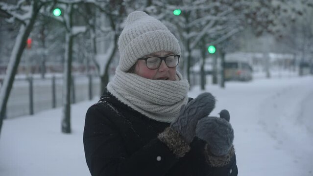 Woman in warm clothes and glasses freezes in winter and warms up her hands in mittens. Snowfall in city near road with cars. Winter bad weather and discomfort for shivering person.