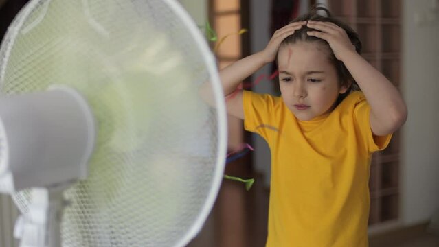Children Suffering From Summer Heat Enjoy Fresh Cool Breeze Blowing Fan. Boy in Front Fan Hot Day on Home in Living Room. Child Enjoying Cool Wind From Electric Fan at Home at Summer Heat Weather.