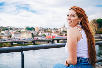 Fototapeta na wymiar woman posing on the bridge. a girl with red hair smiles while sitting on a bridge, a landscape of a town in the background. city atmosphere concept