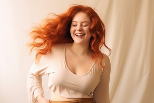 Happy girl. Beautiful young plus size woman with long red hair smiles, laughs, dances on light beige background. Concept of self-love, body positivity. Human emotions