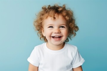 Plump handsome child todler boy with curly hair laughs with his eyes closed in joy in white T-shirt on blue background. Happy baby, childish joy. Concept of people's emotions