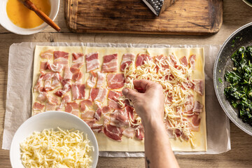 The female cook prepares mini pizza cakes, sprinkles cheese on puff pastry with bacon. - obrazy, fototapety, plakaty