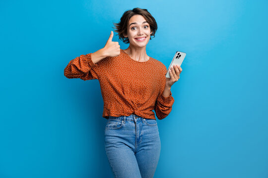 Photo portrait of pretty young girl hold gadget thumb up excited wear trendy brown outfit isolated on blue color background