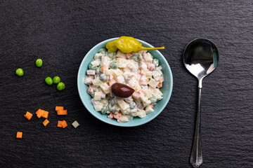 A bowl with the appetizer Ruska Salata, Bulgarian variation of Olivier salad, placed on black stone next to a spoon, spread green peas, diced pickled carrots  and garnished with an olive and pepper