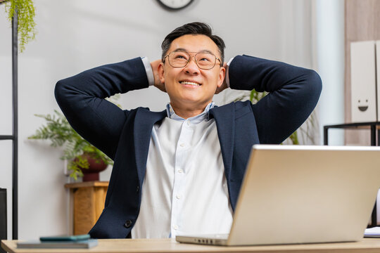 Asian mature businessman freelancer taking break leaning on chair after working celebrating success victory at home office workplace desk. Male Chinese in formal suit puts hands behind head relaxing