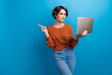 Photo portrait of attractive young woman hold netbook point empty space dressed stylish brown clothes isolated on blue color background