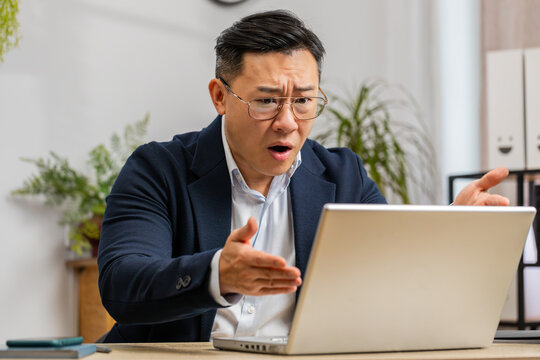 Irritated tired Asian business man while working on laptop, unexpected online website problem, computer virus data loss by hacking. Freelancer feeling mad about broken notebook at office workplace