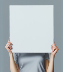 Woman holding blank paper sheet in front of her face. Mock up, blank sheet of paper. Female hands holding blank paper sheet on grey background. Front view mockup with copy space, no face