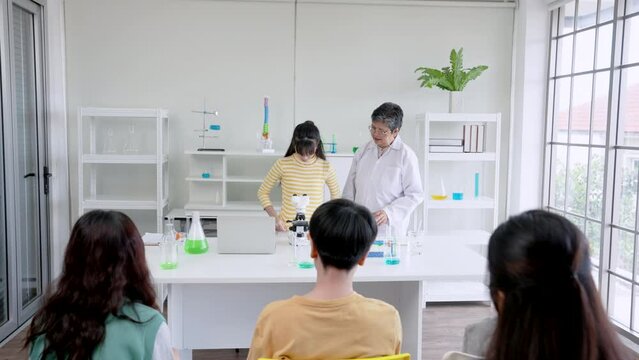 Cute female student with beautiful long hair, standing looking into microscope in front classroom, teacher is standing nearby, girl look through telescope see objects learned set example for friends.