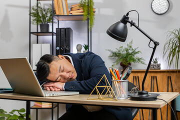 Bored sleepy Asian businessman worker working on laptop computer, leaning on hand falling asleep at...