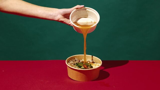 Making delicious ramen. Woman pouring broth onto plate with noodles, vegetables and spices over green red background. Concept of asian food, taste, culinary, pop art style, ingredients.