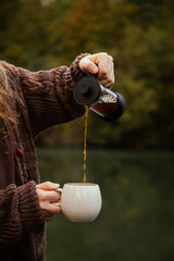Man with long hair pouring coffee in a mug outside in nature by the river