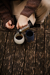 Man with long hair pouring coffee in a mug outside in nature by the river