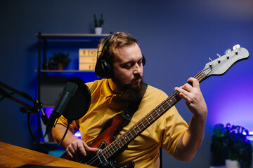 Obraz premium A man in headphones with a microphone on the table plays the bass guitar during an online broadcast. Online media or recording studio concept.
