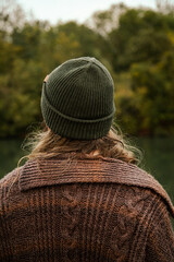 man with long hair in a whool coat and beanie holding a cup of coffee and relaxing looking forward at the river.