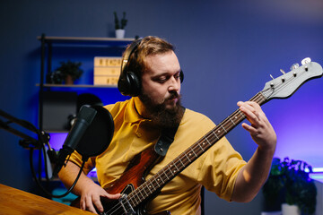 Obraz premium A man in headphones with a microphone on the table plays the bass guitar during an online broadcast. Online media or recording studio concept.
