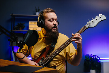 Obraz premium A man in headphones with a microphone on the table plays the bass guitar during an online broadcast. Online media or recording studio concept.