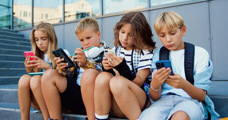 Tenager schoolchildren of two Caucasian girls and boys holding mobile phone and browsing while sitting on stair during school break, oudoors - Powered by Adobe