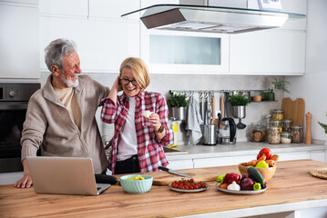 Senior couple cooking dinner together in the kitchen for golden wedding anniversary, reading recipe from internet on laptop. Older man and woman making food for family they invite to celebrate love. - obrazy, fototapety, plakaty