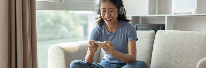 Asian woman playing games on mobile phone and wearing headphones having fun, Play games on the sofa...