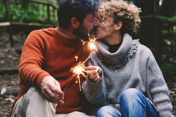 Romantic adult couple kissing in love holding fire sparklers sitting in the nature park enjoy outdoor relationship and leisure activity together. Concept of happy people enjoying vacation - obrazy, fototapety, plakaty