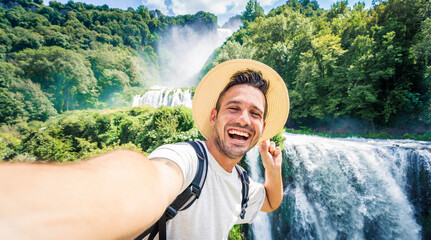 Happy man with hat and backpack taking selfie portrait walking in the forest - Young traveler laughing at camera into the wild - Travel influencer recording social media video content