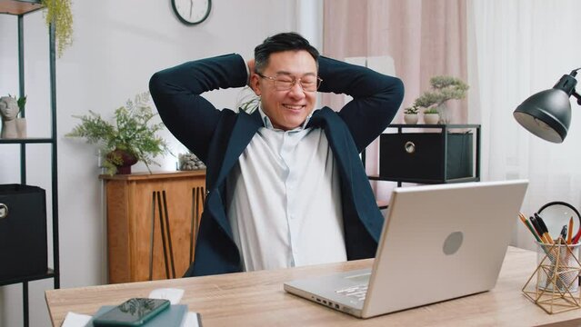 Asian businessman freelancer taking break leaning on chair after working celebrating success victory at modern home office workplace desk. Male Chinese in formal suit puts hands behind head relaxing