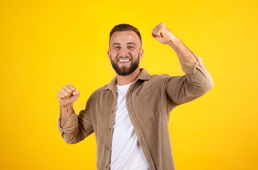 Happy millennial european man with beard in casual has fun, rise fists, celebrate win