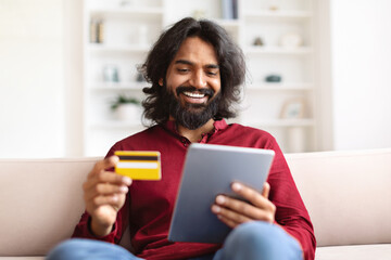 Happy indian guy banking from home, using tablet and card