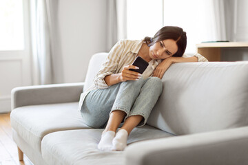 Upset Young Woman Looking At Smartphone Screen, Sitting On Sofa At Home