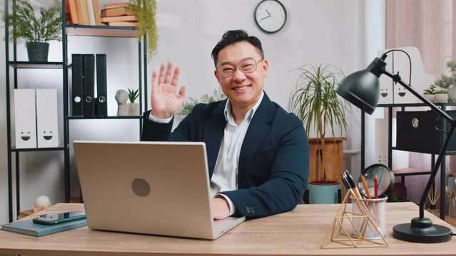 Hello. Asian business man working on laptop computer smiling friendly at camera and waving hands gesturing hi, greeting or goodbye, welcoming with hospitable expression at home office workplace desk