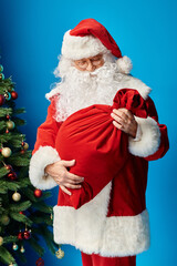Santa Claus with beard and eyeglasses looking at red sack bag with Christmas presents on blue