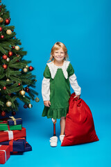 pleased kid in dress with prosthetic leg holding sack bag with presents near Christmas tree on blue