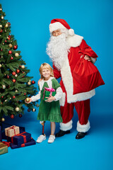 Santa Claus holding sack bag near cheerful girl with prosthetic leg next to Christmas tree on blue