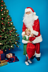 happy girl with prosthetic leg holding sack bag near Santa Claus next to Christmas tree on blue