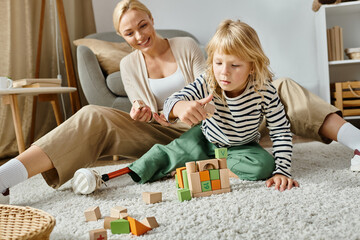 little girl with prosthetic leg sitting on carpet and playing with wooden blocks near happy mother